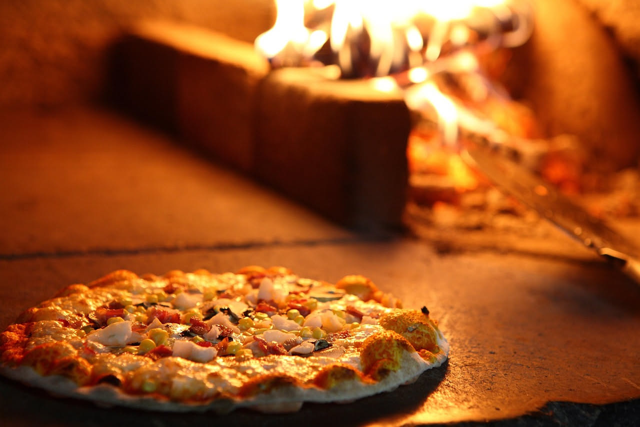 Interno di un forno a cupola tradizionale in mattoni rossi, con fiamme visibili e pizza appena infornata sul fondo, bagliori di calore arancione sulla volta