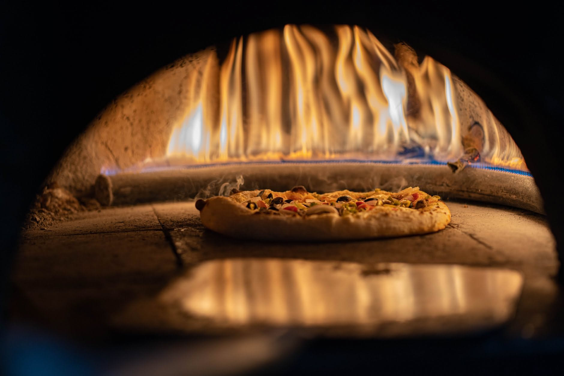 Pizza appena sfornata con bordo dorato su una pala di legno, teglia nera in primo piano, interno di forno domestico sullo sfondo