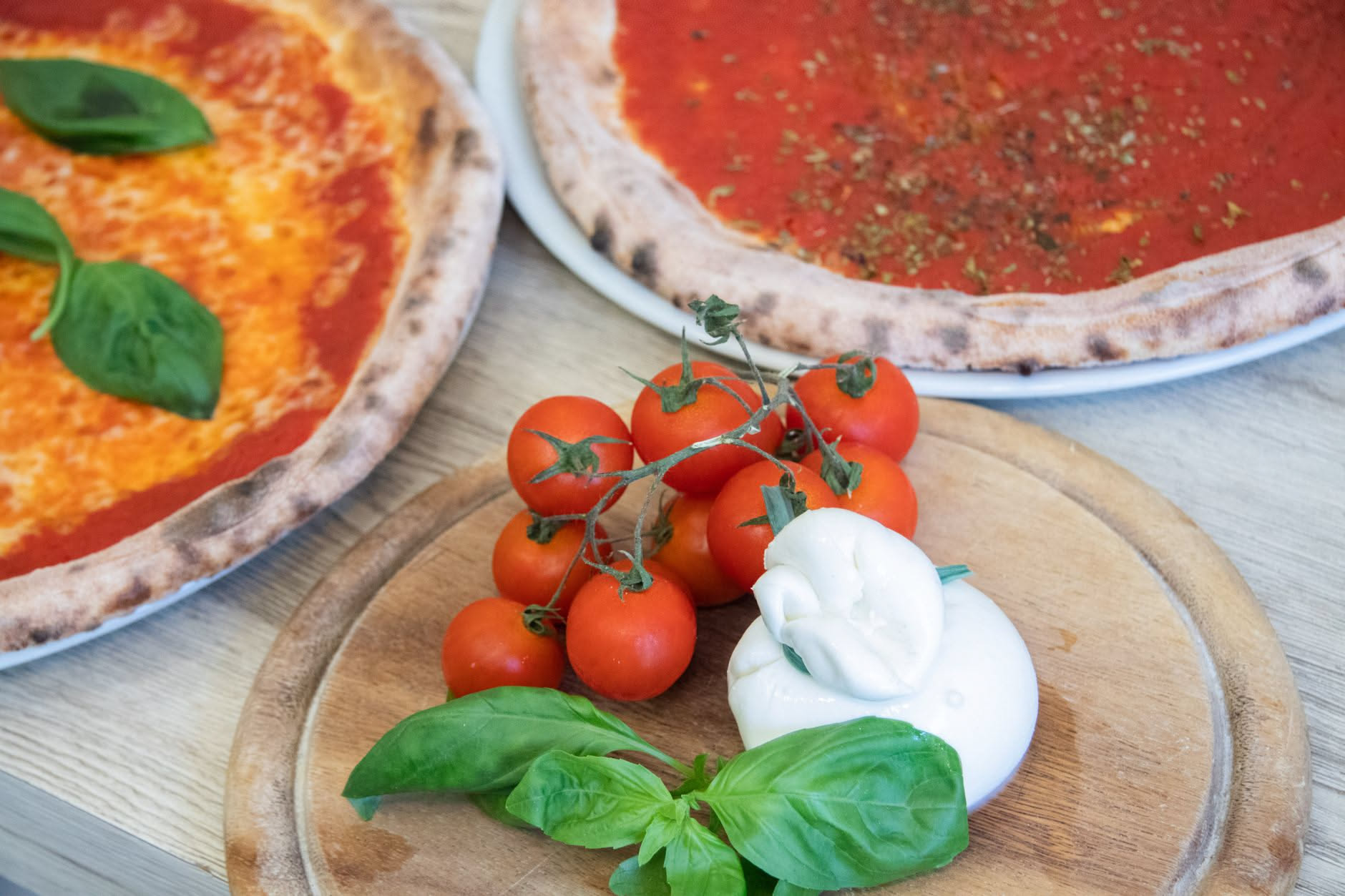 Mano di un pizzaiolo che stende un impasto di pizza su un piano di lavoro in marmo, con forno a legna sullo sfondo sfuocato e fuoco arancione visibile