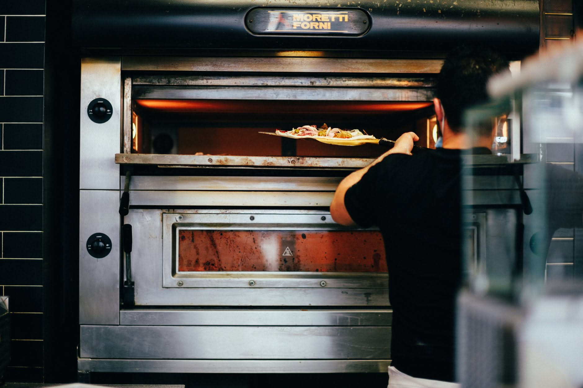 Interno di forno professionale con accesso aperto, mostrando la camera di cottura con pareti refrattarie chiare e una pizza in cottura